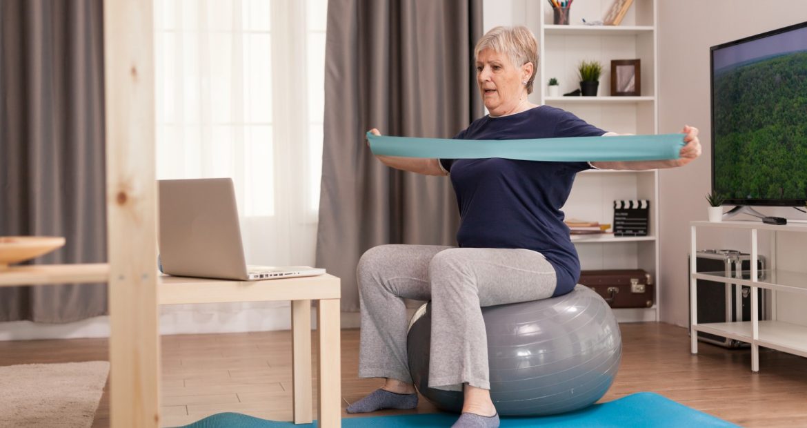 Elderly woman workout at home in front of the laptop. Old person pensioner online internet exercise training at home sport activity with dumbbell, resistance band, swiss ball at elderly retirement age